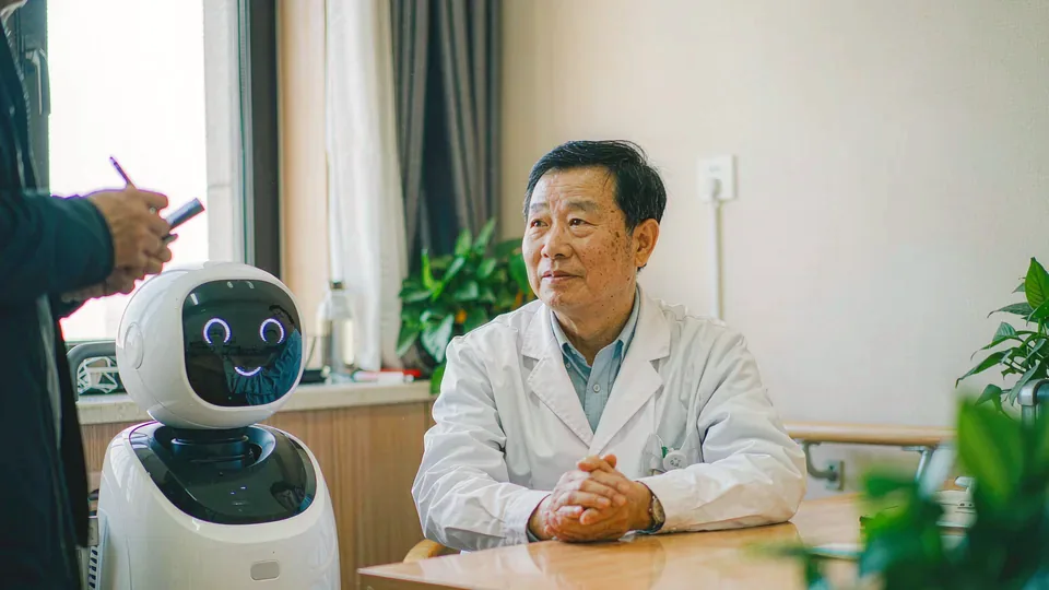 医院康复科会客角：一位穿白大褂的主任医师坐在桌后受访，采访者背影在前景，桌上立着一台白色服务机器人