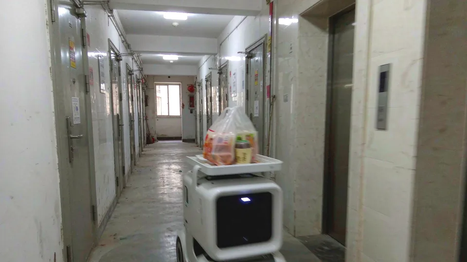 住宅楼道里，配送机器人托着一袋外卖往前走，旁边是防火门与电梯口